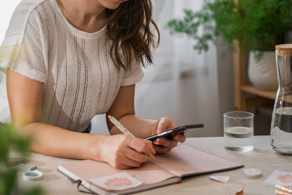 woman writing in notebook while looking at phone